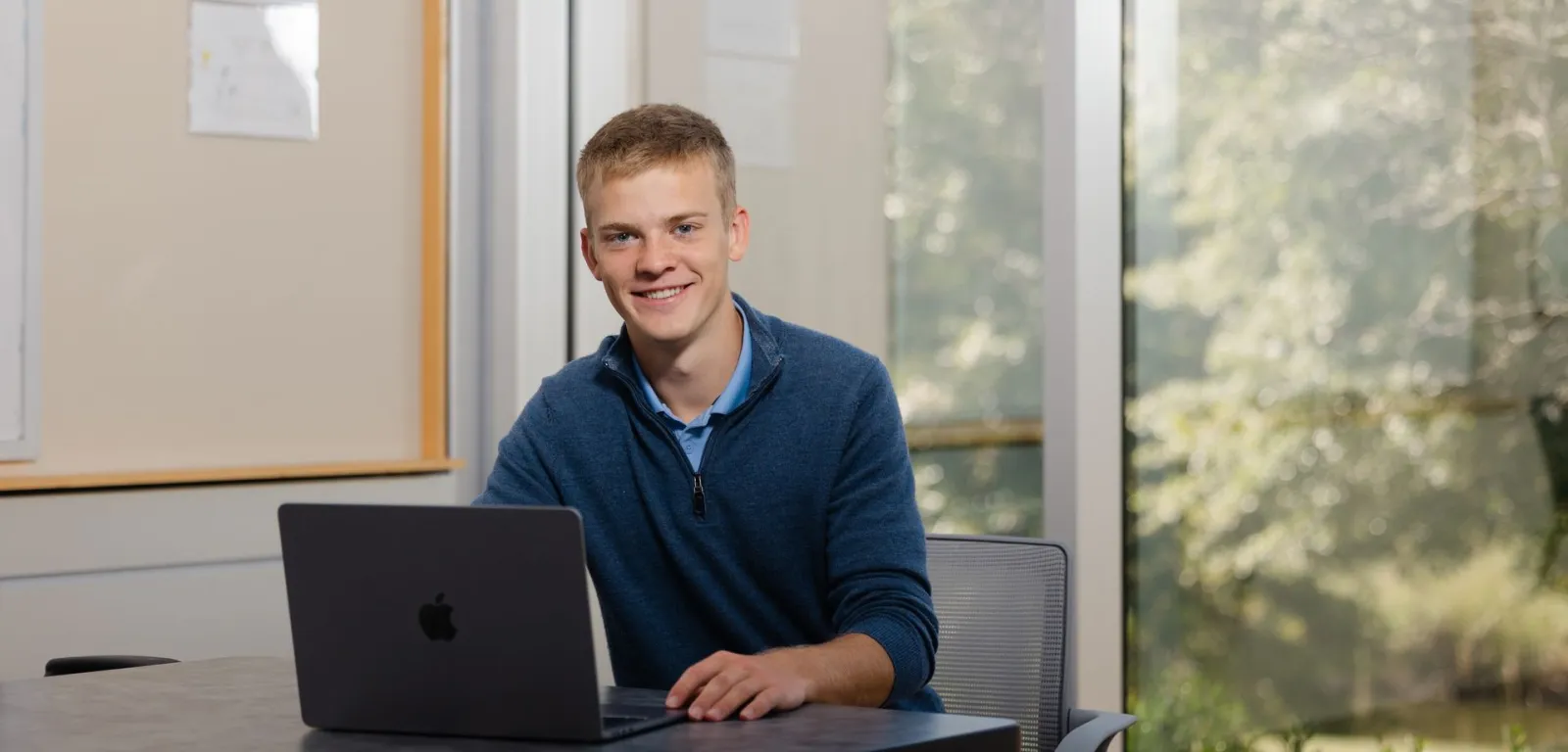 Jared Swift smiling at a table with his laptop, trees outside the window behind him